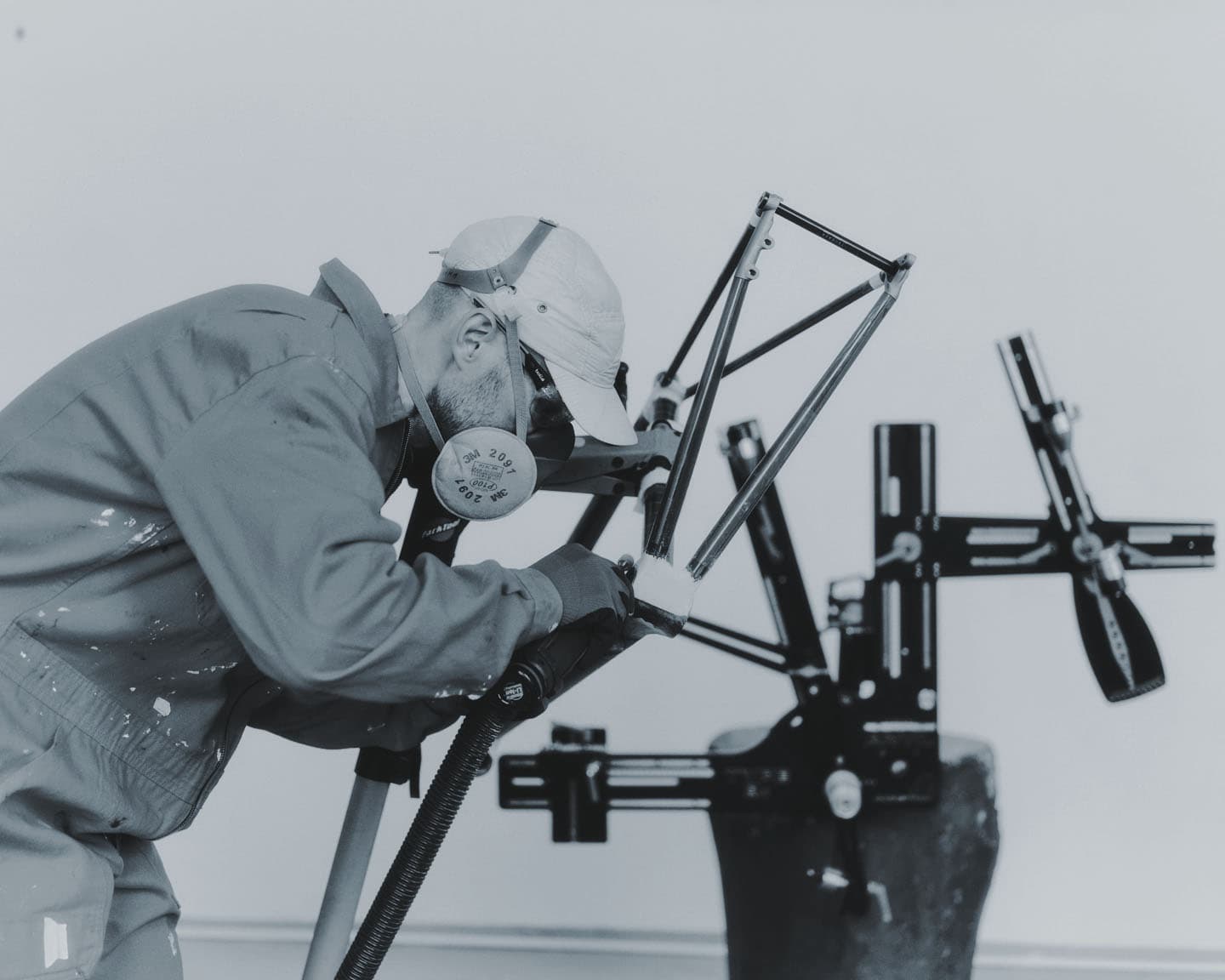 A Tsubasa builder in a respirator and cap, bent over a frame held in a jig, sanding it in the studio