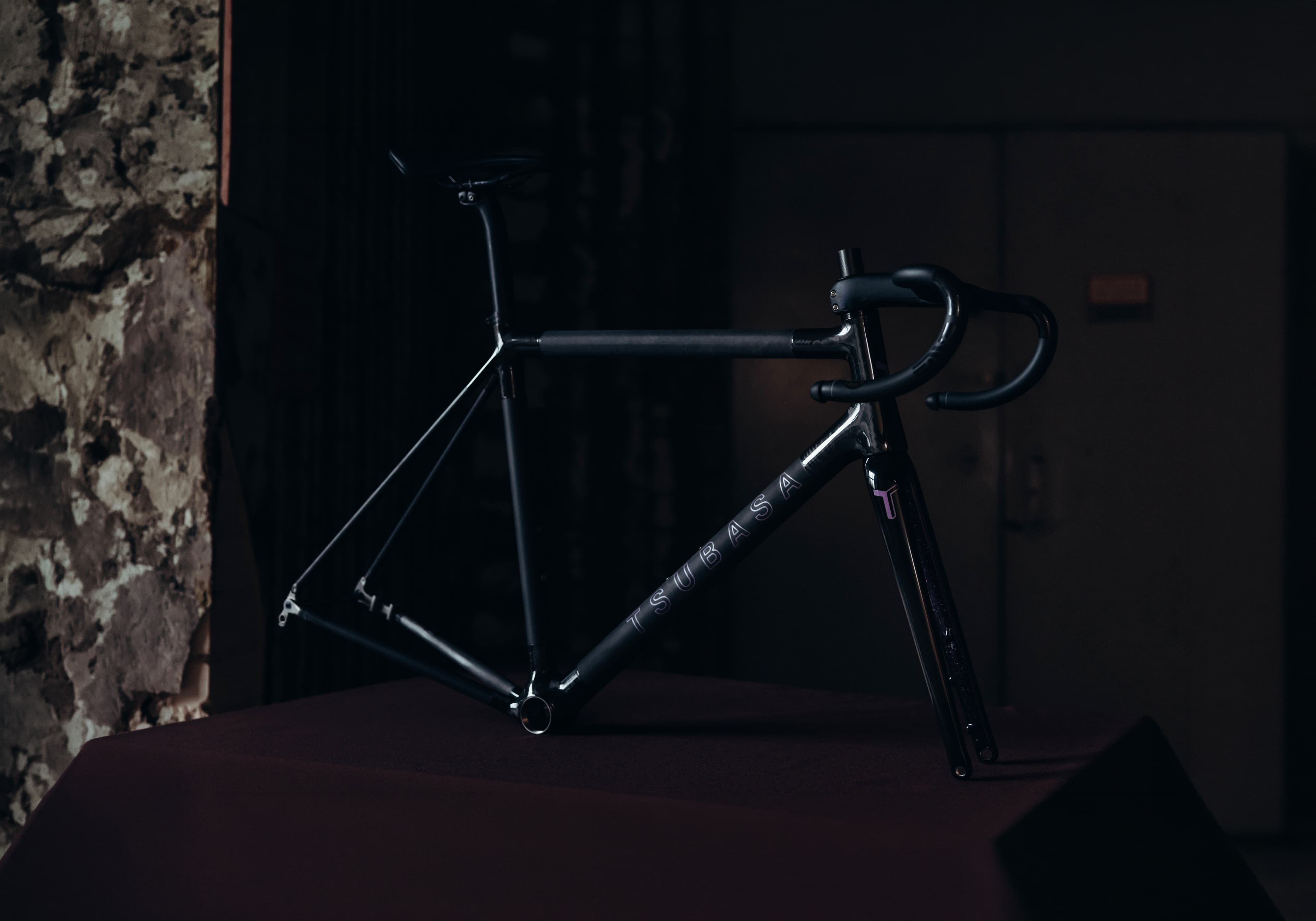 A black Tsubasa frameset on a dark table in a low-lit room, a peeling plaster wall to one side
