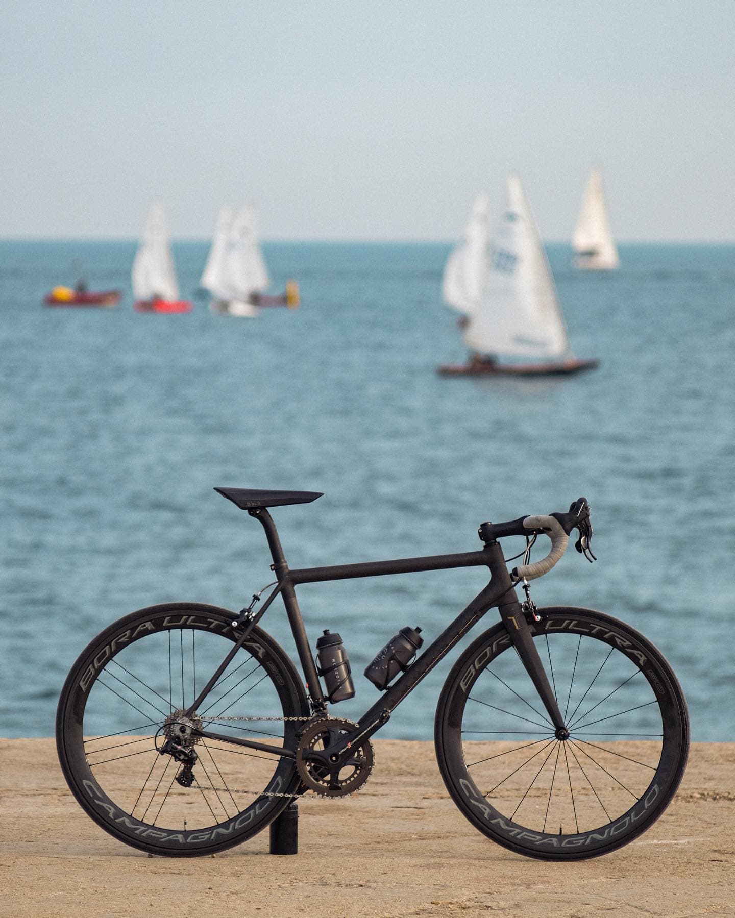 A bike on the sand at the shore, sailing boats out on the water behind